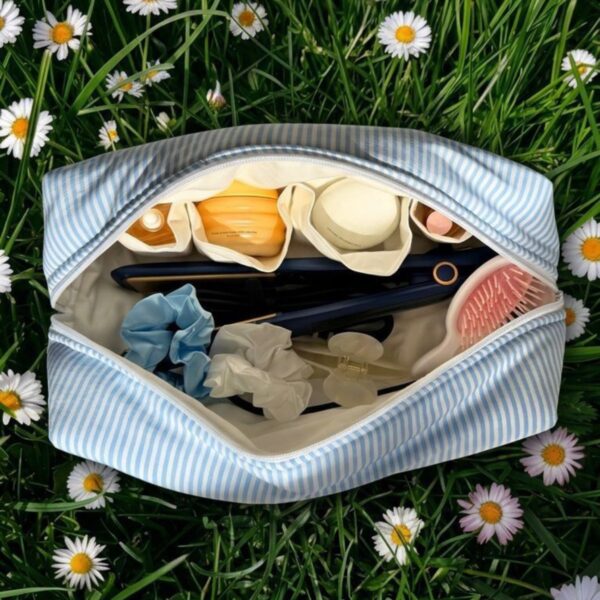 Open lichtblauw-wit gestreepte La Lunore Breeze toilettas met cosmetica en haarstylingtools, op gras met madeliefjes.