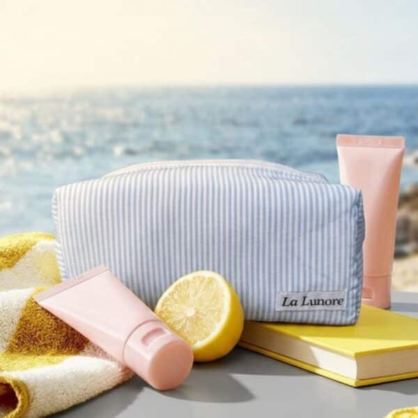 Lichtblauw gestreepte La Lunore toilettas naast een handdoek, citroen en twee roze tubes op een tafel aan zee.