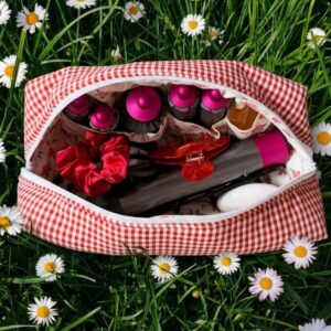Open rood-wit geruite La Lunore Cherry toilettas met stylingtools, scrunchie en accessoires op kunstgras met madeliefjes.