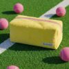 Gele La Lunore toilettas met roze rits op groen kunstgras met een witte lijn en roze tennisballen.